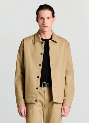 Beiger Blazer aus leichtem Stoff, mit Kragen, schwarzen Knöpfen und lockerem Schnitt, getragen über einem schwarzen Oberteil und einer schwarzen Hose.