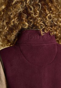 Burgunderfarbener Fleece-Weste mit hohem Kragen, geprägt mit dem Text "JOULES", und sandfarbenen Stoffakzenten. Weicher Stoff mit enger Passform.
