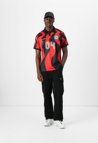 Red and black patterned polo shirt with short sleeves, featuring a collar and number "04." Paired with black cargo pants and white sneakers.