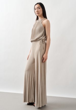 Nő bézs pliszírozott ujjatlan felsőben és bőszárú nadrágban áll, enyhén balra néz egy egyszerű fehér háttér előtt.