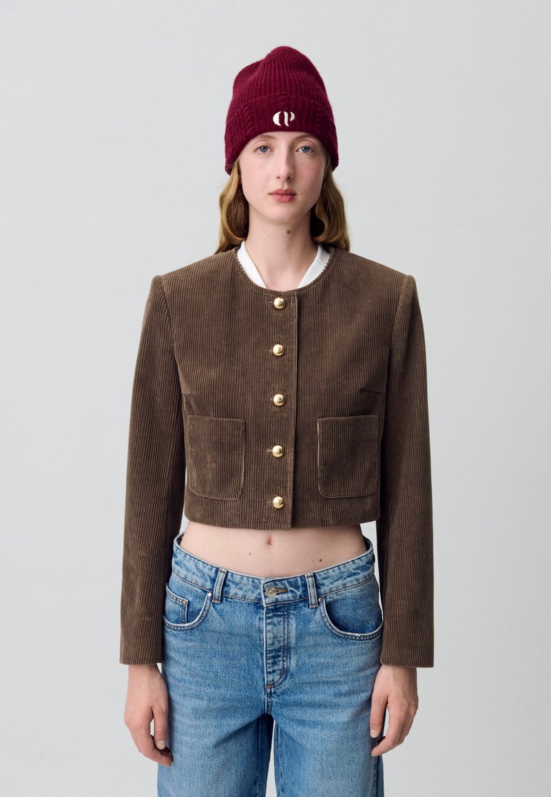 Giacca corta in corda marrone con maniche lunghe, bottoni dorati e due tasche frontali, abbinata a jeans blu a vita alta e a un berretto bordeaux.