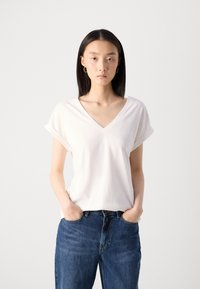 T-shirt blanc à col en V avec des manches courtes retroussées, texture douce, coupe décontractée, associé à un jean taille haute bleu foncé, les mains dans les poches.