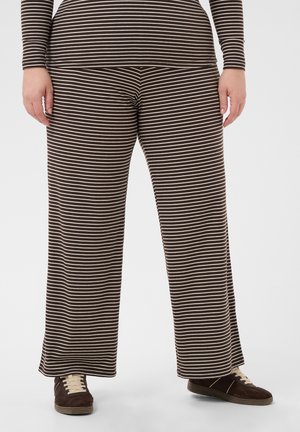 Personne portant un pantalon large rayé horizontalement marron et blanc ainsi qu'un haut assorti, debout avec des baskets marron sur fond blanc.