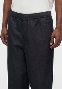 Gros plan sur un pantalon en denim foncé avec une taille élastique porté par une personne avec les mains reposant près des hanches et une chemise blanche rentrée.