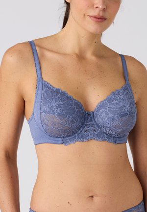 Vrouw die een blauwe kanten bh draagt met bloemmotieven en verstelbare bandjes, van schouders tot taille weergegeven tegen een effen achtergrond.