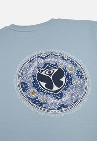 Lichtblauw t-shirt met een cirkelvormig, mozaïekachtig ontwerp met een donkerblauw embleem in het midden, omringd door tekst en kleurrijke stippen.