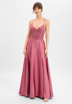 Femme portant une robe longue couleur rose poudré avec un corsage orné de perles, des bretelles fines et une jupe en satin fluide sur un fond blanc uni.