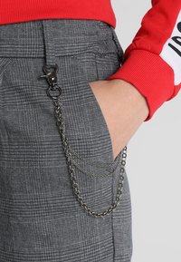 Grå rutiga byxor med en sidficka som har detalj av silverkedja och viklås. Röd långärmad skjorta synlig vid handleden.