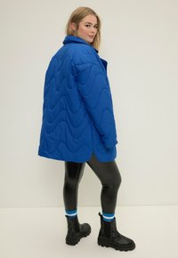 Giacca blu trapuntata con motivo a onde, vestibilità oversize, leggings in ecopelle neri e stivali neri alla caviglia con suola testurizzata e calzini blu a contrasto.
