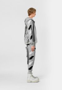 Joven con sudadera con capucha y pantalones grises con estampados de alas en blanco y negro, llevando botas blancas gruesas con cordones, de pie de lado sobre fondo blanco.