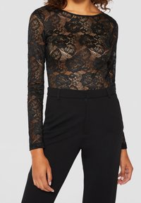Black lace long-sleeve top with floral pattern, semi-sheer fabric, round neckline, paired with high-waisted black trousers.