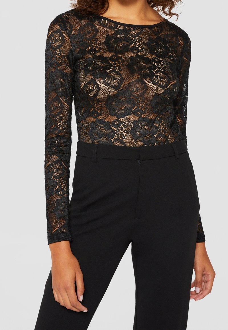 Black lace long-sleeve top with floral pattern, semi-sheer fabric, round neckline, paired with high-waisted black trousers.