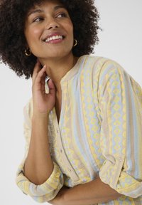 Chemise légère à motifs dans des tons de jaune et de bleu avec une fermeture à boutons, des manches retroussées et un tissu texturé présentant des designs géométriques.