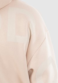 Sweatshirt rose clair avec une texture douce, présentant de grands motifs de logo discrets dans une teinte légèrement plus foncée. Coupe ample avec un col montant.