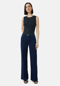 Donkergrijze mouwloze top met een textuurpatroon, gecombineerd met hoge taille donkerblauwe wijde jeans. Model staat tegen een witte achtergrond.