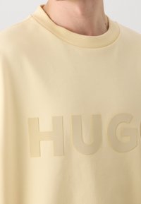 Sweat-shirt beige à col rond avec des lettres en relief formant le mot « HUG » sur la poitrine, porté par une personne dont le visage est cadré de l'arête du nez vers le bas.