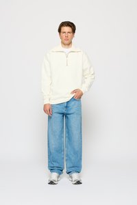 Sudadera cremosa con cremallera y cuello alto, combinada con jeans de mezclilla azul claro holgados y zapatillas blancas, contra un fondo claro y sencillo.