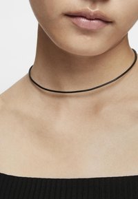 Collana choker nera realizzata in pelle sottile, posata sulla pelle nuda, con un design liscio e minimalista privo di chiusure o decorazioni visibili.