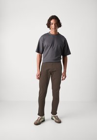 T-shirt gris à manches courtes, pantalon marron fuselé et sneakers brun-vert. Texture du tissu lisse avec un style décontracté. Fond uni.