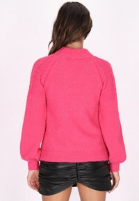 Flauschiger rosa Pulli mit hohem Kragen, Raglanärmeln und Zopfmuster-Details. Gerippte Bündchen und Saum, über einem schwarzen Minirock getragen.
