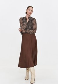 Luźna brązowa spódnica plisowana w połączeniu z czarną bluzką w kwiatowy wzór, z wysokim dekoltem i długimi rękawami, uzupełniona beżowymi kozakami do kolan.