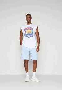 Άνδρας που στέκεται φορώντας αμάνικο λευκό μπλουζάκι Los Angeles Dodgers World Series Champions, ανοιχτό μπλε σορτς, λευκές κάλτσες και λευκά αθλητικά παπούτσια.