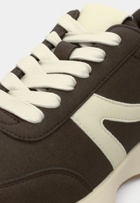 Gros plan d'une basket marron avec des lacets blancs épais et un motif blanc incurvé sur le côté.