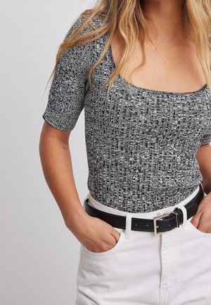 Mujer con un top tejidos con textura en blanco y negro, mangas hasta el codo, metido dentro de pantalones blancos con cinturón negro, con las manos en los bolsillos.