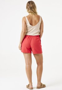 Pantalones cortos rojos con una banda elástica en la cintura, combinados con una blusa sin mangas color beige con un escote bajo en la espalda. Llevado con sandalias marrón claro.
