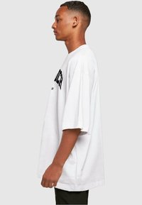 Bílý oversized tričko s kulatým výstřihem a krátkými rukávy, na přední straně s černým potiskem. Model zobrazen z boku.