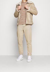 Beige jacka med skinnfoder, ljusbrun stickad tröja, ljusbeige byxor, vita sneakers. Enkel design med en avslappnad passform.