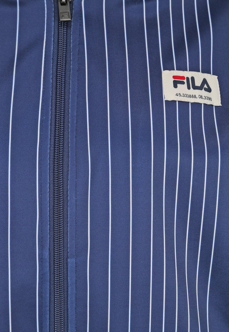 Giacca zip-up blu navy con strisce bianche verticali, caratterizzata da una texture liscia e un'etichetta con il logo FILA sul lato sinistro inferiore. Dettaglio della cerniera visibile.