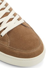 Ruskean mokkasaappaat, joissa on kermaiset nauhat, pyöreä kärki ja teksturoitu pinnan rakenne. Pohja on vaaleanvalkoinen ja siinä on ompelusyksityiskohtia reunassa.