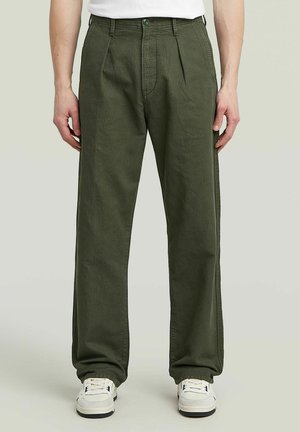 Pantalons larges pour hommes en coton vert olive avec plis à l'avant, dotés d'une fermeture à bouton et de poches latérales, associés à des baskets blanches.