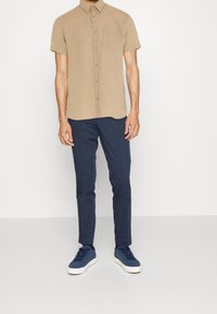 Camisa corta de manga corta en color beige con un bolsillo en el pecho, combinada con pantalones ajustados en azul marino y zapatillas azul marino con suelas blancas.