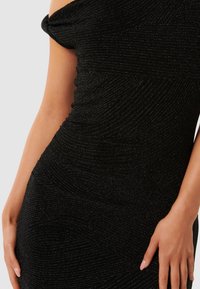 Rochie neagră cu umerii descoperiți, din material texturat, cu un model ondulat. Imaginea de aproape arată țesătura netedă și strălucitoare și silueta ajustată.