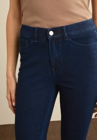 Jeans din denim albastru închis, cu un design ajustat, buzunare, închidere cu buton și o textură netedă, purtate cu un top bej.