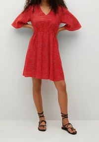 Robe rouge avec un décolleté en V, des manches courtes bouffantes et une taille élastique. Caractérisée par un motif de points blancs épars et une jupe évasée.