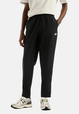 Personne portant un pantalon de survêtement noir New Balance avec une taille élastique et des baskets blanches, debout les mains dans les poches.