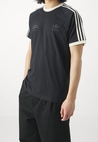 Osoba oblečená v černém tričku Adidas Originals s bílými pruhy na rukávech a černých kalhotách, stojící na bílém pozadí.