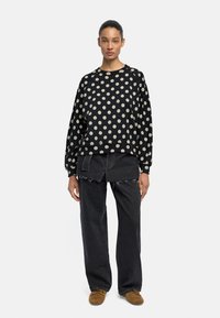 Czarna bluza w kropki z prążkowanymi mankietami i dołem, zestawiona z ciemnymi dżinsami z postrzępionymi krawędziami i warstwowym krojem. Brązowe buty.