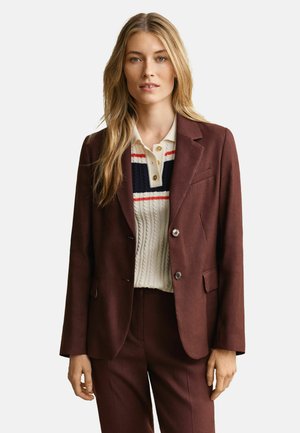 Femme blonde portant un costume marron composé d'une veste et d'un pantalon, avec un pull en maille torsadée blanc orné de rayures rouges et bleu marine.