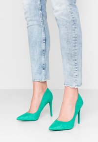 Pompes à talons hauts en velours vert avec des bouts pointus. Les chaussures présentent un design épuré et une texture lisse, associées à un jean court bleu clair.