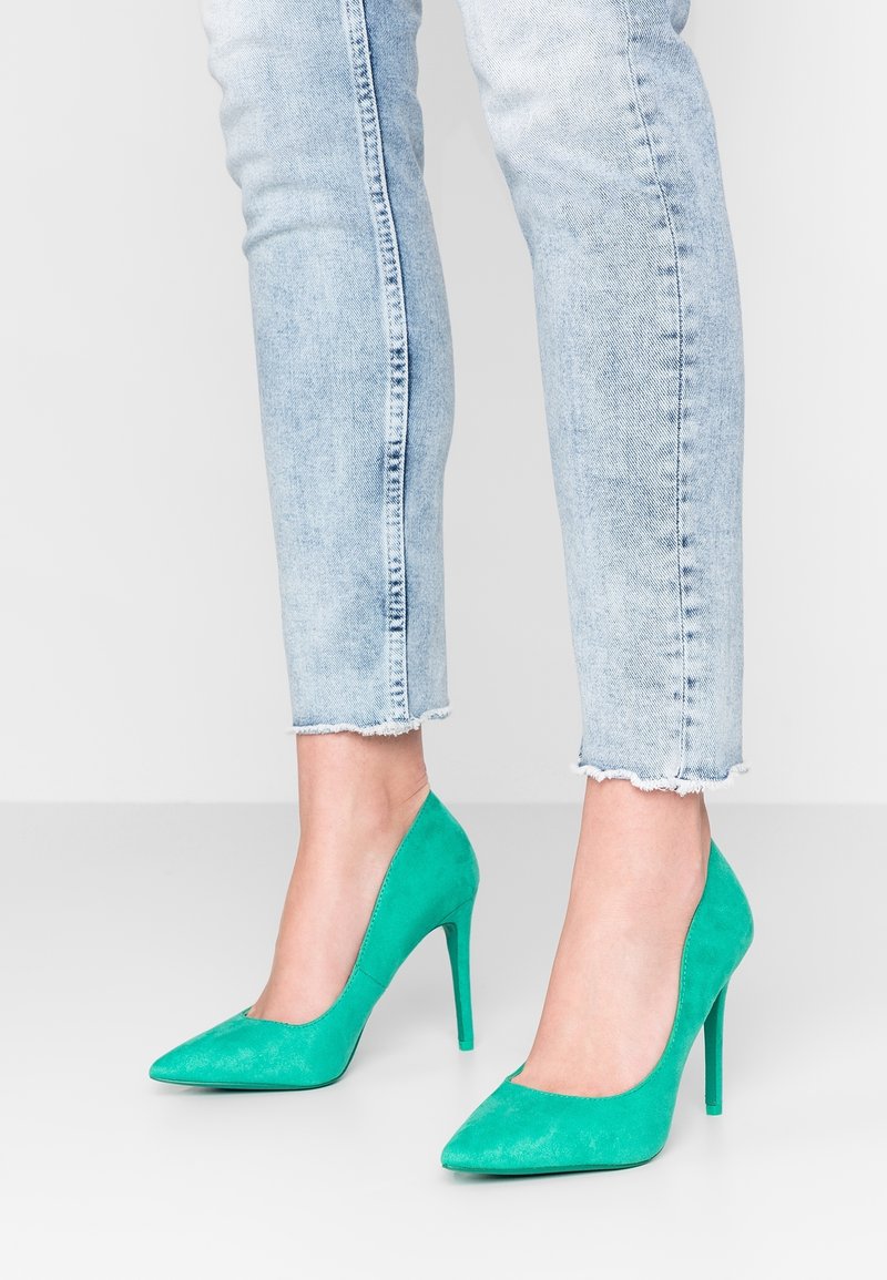Pompes à talons hauts en velours vert avec des bouts pointus. Les chaussures présentent un design épuré et une texture lisse, associées à un jean court bleu clair.
