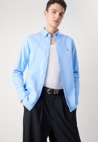 Camicia azzurra chiara a maniche lunghe in cotone liscio, indossata sopra una canotta bianca, abbinata a pantaloni neri a righe e una cintura nera.
