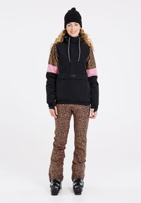 Schwarze Ski-Jacke mit Ärmeln im Leopardenmuster und pinken Akzenten, kombiniert mit einer Hose im Leopardenmuster. Das Modell trägt eine schwarze Beanie und Skischuhe.