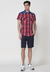 Kurzarm-rot-karierte Hemd mit blauen Jeans-Schultern, zwei Brusttaschen, kombiniert mit navy-blauen umgeschlagenen Shorts und grauen Sneakers.