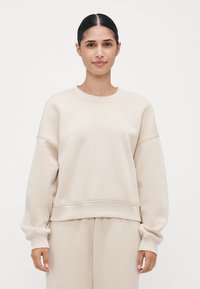 Ung kvinne med mørkt hår i hestehale, iført en kremfarget oversized hettegenser og matchende bukser foran en enkel hvit bakgrunn.
