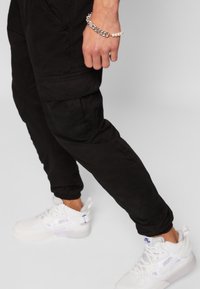 Zwarte cargobroek met elastische boorden, meerdere zakken en een losse pasvorm. Gecombineerd met witte sneakers met een gestructureerd ontwerp en logo.