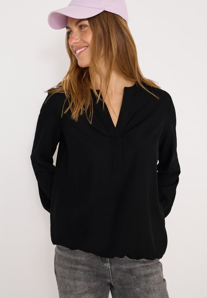 Schwarze Bluse mit langen Ärmeln und V-Ausschnitt aus weichem Stoff. Kombiniert mit hellgrauen Jeans und einer lavendelfarbenen Baseballkappe. Glatte Textur.